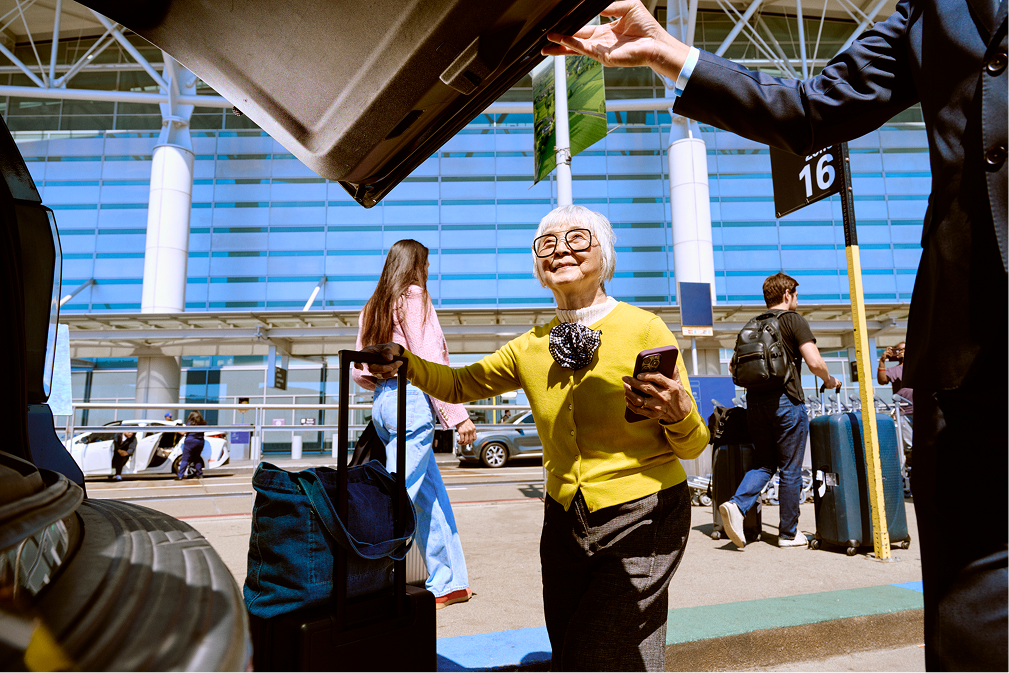 Older woman in yellow sweater with luggage at airport curb, holding phone, car trunk open.